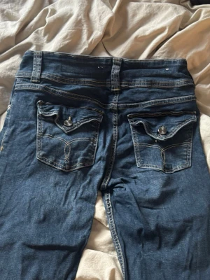 Blå jeans med ficklock och knappar - Säljer ett par klassiska blå jeans med raka ben och snygga detaljerade bakfickor med ficklock och knappar. Jeansen har normalhög midja, bälteshällor och kontrastsömmar. Perfekta till vardagslooken och passar till det mesta.