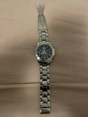 Silverfärgad klocka från Hugo Boss - Stilren klocka från Hugo Boss med silverfärgad länkarmband i metall och mörkblå urtavla. Klockan har kronograf-funktion med tre mindre urtavlor och tydliga indexmarkeringar. Klassisk design som passar till många olika stilar. Behöver fixas 