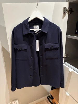 Mörkblå overshirt från Lindbergh - Stilren mörkblå overshirt från Lindbergh i relaxed fit. Tillverkad i superflex-material. Två bröstfickor med lock och knapp, klassisk krage och knäppning framtill. Perfekt lager på lager-plagg för en modern look. Helt ny aldrig använd.
