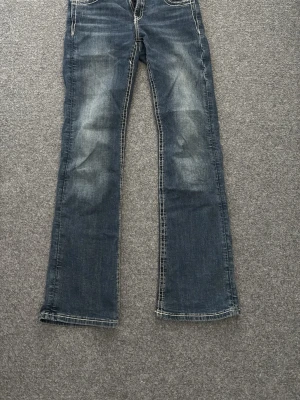 Blå bootcut jeans med broderade fickor - Säljer ett par blå bootcut jeans med slitningar framtill och snygga broderade detaljer på bakfickorna. Jeansen har kontrastsömmar och klassisk fyraficksmodell. Perfekta för dig som gillar en avslappnad men ändå trendig stil.
