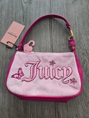 Rosa Juicy Couture handväska - Söt rosa handväska från Juicy Couture med broderad text 'Juicy' och fjäril på framsidan. Väskan har en axelrem i mörkrosa, guldfärgade detaljer och dragkedja med hjärtformad berlock. Materialet är en mix av mocka och konstläder för en trendig look.