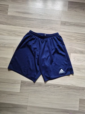 Marinblå träningsshorts från Adidas - Säljer ett par marinblå träningsshorts från Adidas med elastisk midja och klassisk logga på ena benet. Shortsen är tillverkade i ett lätt och svalt polyestermaterial, perfekta för sport och aktivitet.