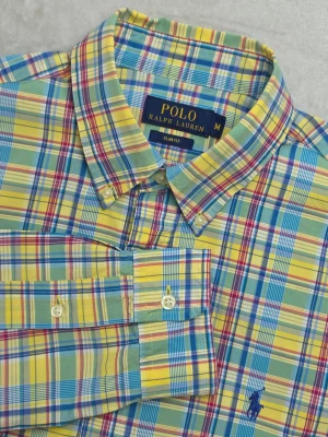 Polo Ralph Lauren Slim Fit skjorta rutig flerfärgad storlek M - Rutig skjorta från Polo Ralph Lauren i Slim Fit-passform. Färgstarkt rutmönster i gult, grönt, blått och rött med klassisk preppy-stil. Tillverkad i 100% bomull och försedd med button down-krage samt marinblå broderad Polo-logotyp på bröstet.  Storlek M Slim Fit  Mycket fint skick.  Mått • Bröstbredd: ca 52 cm • Plagglängd: ca 73 cm • Axelbredd: ca 45 cm • Ärmlängd: ca 62 cm  Material 100% bomull  Varan är begagnad och säljs i befintligt skick, se bilder för detaljer.