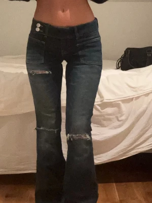 Mörkblå bootcut jeans med slitningar - Säljer ett par mörkblå bootcut jeans med slitna detaljer vid knäna och låret. Jeansen har låg midja, två knappar i linningen och klassiska fickor fram. Materialet är denim med stretch för skön passform. Perfekta för en trendig och avslappnad look.