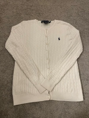 Vit kabelstickad kofta från Polo Ralph Lauren - Kabelstickad kofta i krämvit färg från Polo Ralph Lauren. Klassisk design med rund halsringning, långa ärmar och knäppning framtill. Liten broderad logga på bröstet och ribbade muddar vid ärmslut och nederkant. Perfekt för lager på lager.