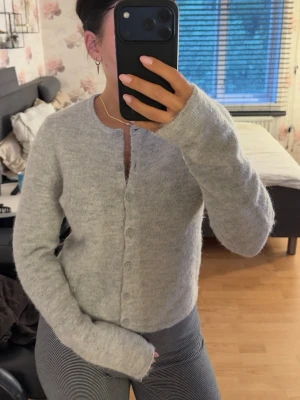 Ljusgrå fluffig kofta med rund hals - Mysig ljusgrå kofta med rund halsringning och knappar framtill. Koftan är fluffig och har långa ärmar med en något croppad passform. Perfekt att slänga på sig för en chill look.