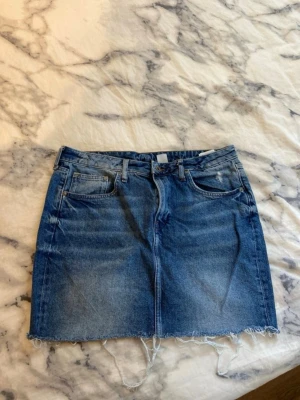 Kort jeanskjol  - Säljer en klassisk blå jeanskjol från &Denim med rå, fransig kant nertill. Kjolen har fem fickor, bälteshällor och stängs med knapp och dragkedja. Perfekt för dig som gillar en avslappnad och trendig stil. Liten i storleken. Passar M 