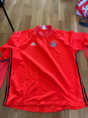 Röd Bayern München träningströja Adidas - Säljer en röd långärmad träningströja från Adidas med Bayern München-logga på bröstet. Tröjan har svarta detaljer längs sidorna och är tillverkad i funktionsmaterialet Climacool som andas. Perfekt för fotbollsträning eller gymmet.