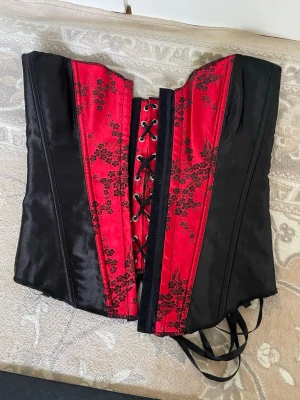 Svart och röd korsett med spetsdetaljer - Snygg korsett i svart och rött med blommiga spetsdetaljer och snörning både fram och bak. Materialet är glansigt och ger en lyxig känsla. Perfekt för dig som vill ha en statement piece till din outfit. Justerbar passform tack vare snörningen.