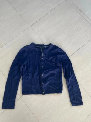 Mörkblå cardigan med knappar - Mysig mörkblå cardigan med rund halsringning och långa ärmar. Tröjan har knäppning framtill med flera knappar och en diskret detalj vid bröstet. Perfekt att slänga över en topp för en enkel och stilren look.