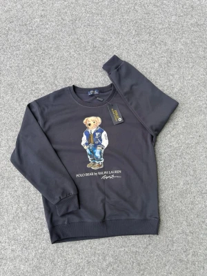 Mörkblå sweatshirt Polo Bear Ralph Lauren - Mörkblå sweatshirt från Ralph Lauren med ikoniska Polo Bear-trycket framtill. Rund halsringning, ribbade muddar och avslut. Skön bomullsmix och avslappnad passform. Perfekt för en casual och trendig look.
