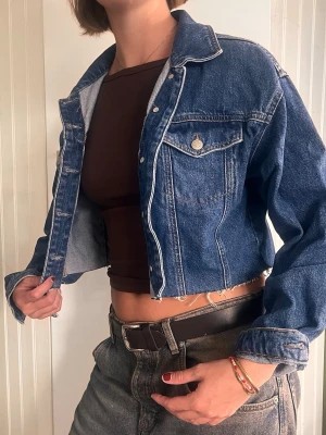 Cropped blå jeansjacka - Trendig cropped jeansjacka i klassisk blå denim med silverfärgade knappar och två bröstfickor med lock. Jackan har en avslappnad passform.
