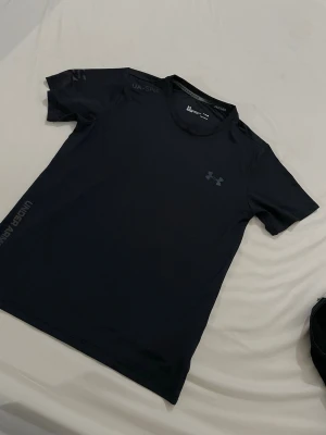 Under armour - Snygg svart tränings-tshirt från Under Armour med reflekterande logga och textdetaljer på bröstet och sidan. Tillverkad i ett lätt och snabbtorkande material som passar perfekt till gymmet eller löprundan. Klassisk passform med rund hals och korta ärmar.