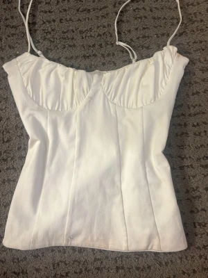 Vit korsettopp med tunna axelband - Säljer en stilren vit korsettopp med tunna justerbara axelband och lätt rynkade kupor. Toppen har en figurnära passform och är perfekt för att skapa en trendig look. Materialet känns mjukt och har en lätt struktur.