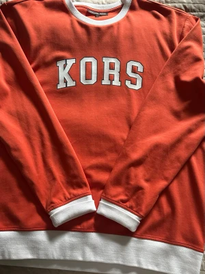 Orange sweatshirt med vit text KORS - Cool orange sweatshirt från Michael Kors med vit text 'KORS' på bröstet. Tröjan har vita ribbade muddar vid ärmslut, halsringning och nederkant. Perfekt för en avslappnad och trendig look. Använd en gång, pris kan diskuteras!