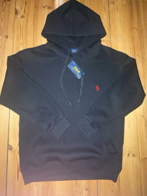 Svart hoodie från Polo Ralph Lauren - Svart hoodie från Polo Ralph Lauren med klassisk huva och dragsnören. Har en röd broderad logga på bröstet och en stor magficka framtill. Tillverkad i mjukt material som känns skönt mot huden. Perfekt för en avslappnad och stilren look. Ser ut som M men den passar X och XS