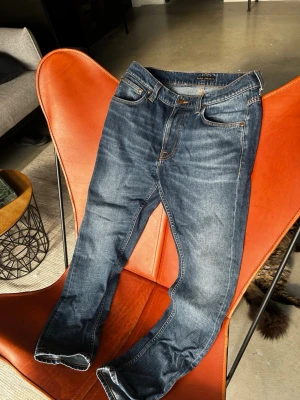 Slitna nudie jeans - Säljer ett par klassiska mörkblå jeans med raka ben och fem fickor. Jeansen har snygga kontrastsömmar och en traditionell knappgylf. Materialet är slitstark denim i bomull och passformen är normal. Perfekta till vardagslooken!