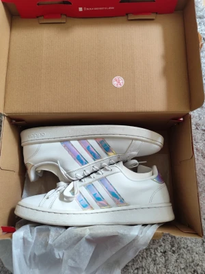 Vita Adidas sneakers med holografiska ränder - Säljer ett par vita Adidas sneakers med holografiska, skimrande ränder på sidorna och matchande detalj på hälen. Klassisk låg modell med snörning och vit sula. Perfekta för dig som vill ha en clean look med lite extra färg.