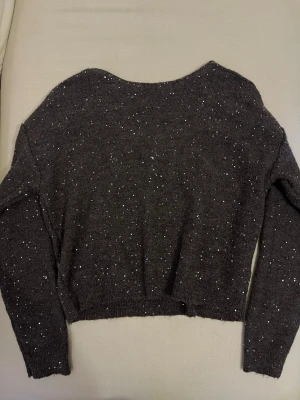 Mörkgrå stickad tröja med glitter - Mysig mörkgrå stickad tröja med långa ärmar och glittriga inslag över hela plagget. Tröjan har en bred, rundad halsringning och är något croppad i modellen. Perfekt för dig som vill ha en enkel men ändå lite extra stickad tröja.
