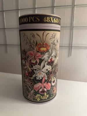 Pussel 1000 bitar med blommotiv - Säljer ett pussel med 1000 bitar i en rund förpackning. Motivet är ett detaljerat blommönster i färger som rosa, vit, grön och gul. Storleken på pusslet är 48x68 cm. Perfekt för dig som gillar utmanande pussel och vackra illustrationer.