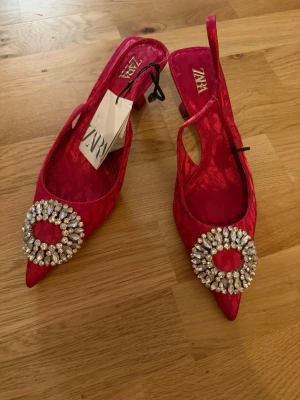 Rosa pumps med strassdetalj från Zara - Säljer ett par snygga rosa pumps från Zara med spetsig tå och öppen häl. Skorna har en stor rund strassdetalj framtill och är tillverkade i ett glansigt tyg med diskret mönster. Perfekta för dig som vill sticka ut med dina skor.