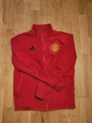 Röd Manchester United ziptröja Adidas - Röd ziptröja från Adidas med Manchester Uniteds klubbmärke broderat på bröstet. Tröjan har hög krage, hel dragkedja och två sidofickor. Tillverkad i mjukt material, perfekt för träning eller chill. Svart Adidas-logga på framsidan.
