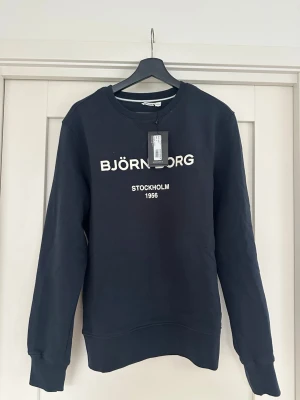 Svart sweatshirt från Björn Borg - Stilren svart sweatshirt från Björn Borg med vit text på bröstet där det står 'BJÖRN BORG STOCKHOLM 1956'. Tröjan har rund halsringning, långa ärmar och ribbade muddar. Perfekt för en avslappnad look.