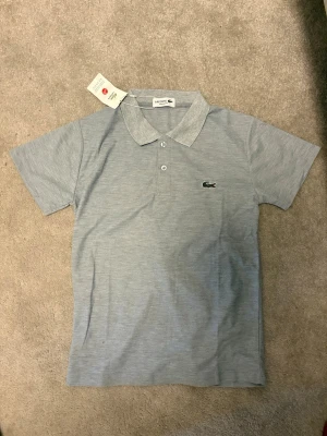 Ljusgrå pikétröja från Lacoste - Snygg ljusgrå pikétröja från Lacoste med klassisk krage och två knappar framtill. Tröjan har korta ärmar och den ikoniska krokodilloggan broderad på bröstet. Perfekt för en clean och stilren look.