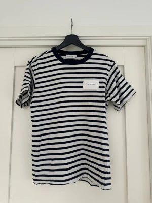 Randig t-shirt från Calvin Klein - Snygg randig t-shirt från Calvin Klein i vitt och marinblått. Klassisk passform med korta ärmar och rund halsringning. Tillverkad i mjuk bomull och har en diskret logga på bröstet. Perfekt för en avslappnad och stilren look.