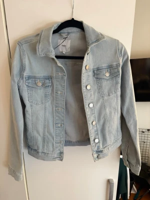 Ljusblå jeansjacka från Next - Snygg ljusblå jeansjacka från Next med klassisk krage, två bröstfickor med lock och silverfärgade knappar. Jackan har raka ärmar och en avslappnad passform som passar perfekt till alla dina outfits.
