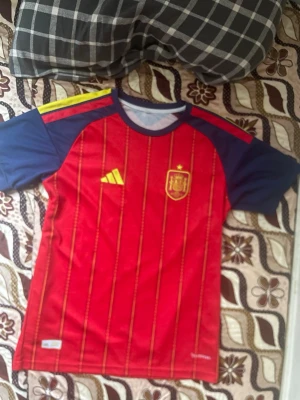Spaniens matchtröja röd/blå - Säljer en officiell matchtröja för Spaniens landslag i rött med blå ärmar och gula detaljer. Tröjan har tunna vertikala ränder, rund halsringning och ett broderat landslagsemblem på bröstet. Tillverkad i ett lätt och ventilerande funktionsmaterial.