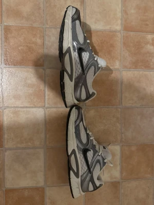 Nike vita och grå sneakers - Nike sneakers i vitt och grått med meshpaneler och detaljer i syntetmaterial. Klassisk låg modell med snörning och svart Nike-logga på plösen. Snygg retrostil som passar till många outfits.