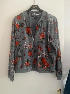 Grå  tunn blommig bomberjacka från ONLY - Snygg grå tunn  bomberjacka från ONLY med färgglatt blommönster i rött, orange och vitt. Jackan har dragkedja framtill, ribbade muddar och två fickor. Perfekt för dig som vill sticka ut med en unik och trendig look.