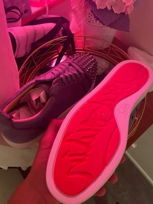 Grå sneakers med nitar och röd sula - Snygga grå sneakers från Christian Louboutin med ikonisk röd sula och silverfärgade nitar på tån. Skorna har en låg profil, snörning och är tillverkade i mocka. Perfekta för dig som vill sticka ut med en edgy och exklusiv stil.