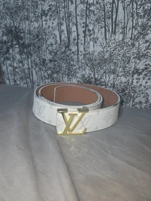 Vitt Louis Vuitton bälte med guldspänne - Snyggt vitt bälte från Louis Vuitton med klassiskt LV-mönster och ett stort guldfärgat LV-spänne. Bältet har en stilren design och insidan är ljusbrun. Perfekt accessoar för att lyfta vilken outfit som helst.