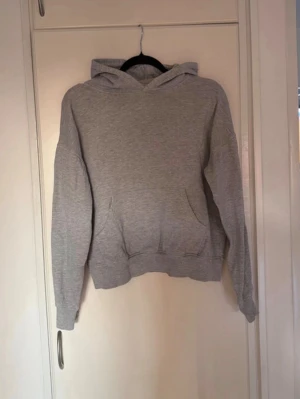 Grå basic hoodie med huva - En enkel och stilren grå hoodie med huva och magficka. Hoodien har långa ärmar, ribbade muddar och är gjord i mjukt sweatshirtmaterial. Perfekt för en avslappnad look och funkar året runt.
