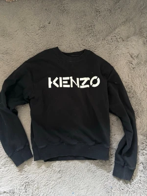 Svart Kenzo sweatshirt med logga - Säljer en svart sweatshirt från Kenzo med stor vit logga framtill. Tröjan har rund halsringning, ribbade muddar vid ärmslut och nederkant samt en avslappnad passform. Perfekt för dig som gillar streetwear och vill ha en stilren look.