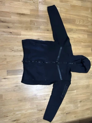 Svart Nike tech med dragkedja - Snygg svart Nike tech med hel dragkedja framtill och reflekterande detaljer. Koftan har en huva, två sidofickor och en extra ficka på ena ärmen. Tillverkad i mjukt material som passar perfekt till streetwear-looken. Om man är lång så passar den M också.