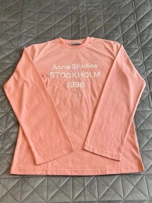 Ljusrosa långärmad topp Acne Studios - Ljusrosa långärmad topp från Acne Studios med tryck 'Acne Studios STOCKHOLM 1996' framtill. Klassisk rund halsringning och relaxed fit. Perfekt för dig som gillar stilrena och trendiga plagg.