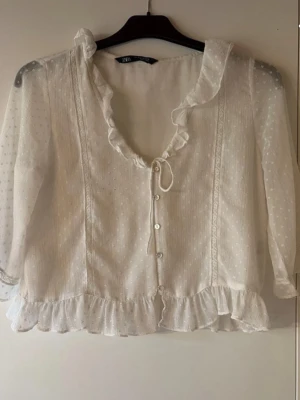 Vit blus med volang från Zara - Söt vit blus från Zara med volangkant vid halsringning och nederkant. Blusen har knappar framtill, diskret prickigt mönster och är tillverkad i ett tunt, transparent tyg. Trekvartsärm och romantisk look som passar perfekt till jeans eller kjol.