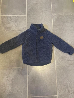 Mörkblå teddyjacka från Ströva - Mysig mörkblå teddyjacka från Ströva med hög krage och dragkedja framtill. Jackan har ett brunt patchmärke på bröstet och är gjord i fluffigt teddy-material som håller dig varm. Perfekt för kyliga dagar och enkel att matcha till olika outfits.
