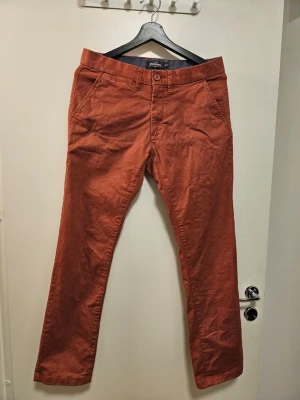 Dressmann terrakottafärgade chinos - Chinos från Dressmann i en varm terrakottafärg. Byxorna har en slim fit-passform och är designade med knapp och dragkedja framtill. De har två snedställda fickor fram och två passpoalfickor med knappar bak.   Endast använda ett fåtal gånger.   Nytvättade med parfymfritt tvättmedel.