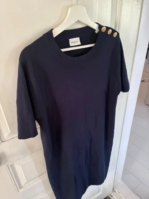 Mörkblå klänning från Redgreen - Mörkblå stickad klänning från Redgreen med rund halsringning och tre dekorativa guldknappar på ena axeln. Klänningen har halvkort ärm och rak passform. Perfekt för en stilren och enkel look.