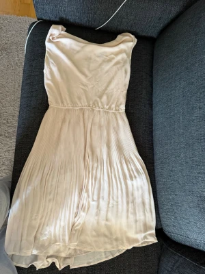 Ljusbeige plisserad klänning - Säljer en ljusbeige klänning med plisserad kjol och enkel, ärmlös överdel. Klänningen har rund halsringning och är tillverkad i ett glansigt, mjukt tyg som ger ett elegant intryck. Perfekt för dig som gillar stilrena och feminina plagg.