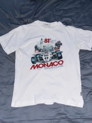 Vit Replay Monaco Grand Prix t-shirt - Säljer en vit Replay t-shirt med tryck av Monaco Grand Prix och en Formel 1-bil på framsidan. T-shirten har korta ärmar och rund hals. Texten 'MONACO REPLAY CORSE' i rött och detaljer i blått och grått. Perfekt för motorsportfans!