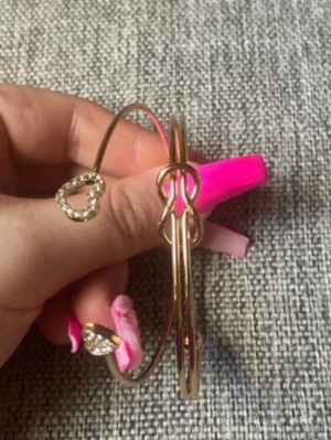 Guldiga armband med hjärtan - Två tunna guldiga armband i metall med öppningsbar design. Ena armbandet har hjärtformade detaljer dekorerade med små glittrande stenar, vilket ger en söt och trendig look. Perfekt accessoar för att lyfta din outfit med lite extra bling.