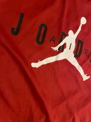 Röd Jordan t-shirt med logga - Snygg röd t-shirt från Jordan med den klassiska Jumpman-loggan i vitt och svart text på bröstet. T-shirten har rund hals och korta ärmar, perfekt för en sportig och avslappnad stil. Tillverkad i mjukt material som känns skönt mot huden.
