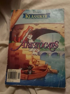 Disney Klassiker Aristocats bok - En färgglad bok ur Disney Klassiker-serien med titeln Aristocats. Omslaget visar de ikoniska katterna vid en bro i Paris och boken har en rosa baksida med text och en tecknad katt. Perfekt för dig som gillar klassiska Disney-berättelser.