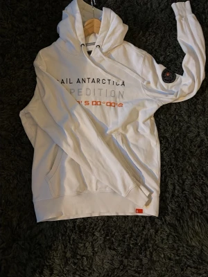 Vit hoodie med tryck och patch - Säljer en vit hoodie med texten 'SAIL ANTARCTICA EXPEDITION' tryckt på bröstet och en patch på ena ärmen. Hoodien har magficka, ribbade muddar och dragsko i huvan. Perfekt för dig som gillar clean stil med detaljer.
