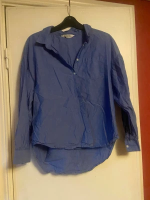 Blå långärmad skjorta i bomull(barn)  - Klassisk blå skjorta i bomull med lång ärm och knappar framtill. Skjortan har en bröstficka och en avslappnad passform med rundad nederkant. Perfekt att styla till jeans eller chinos för en enkel och snygg look.